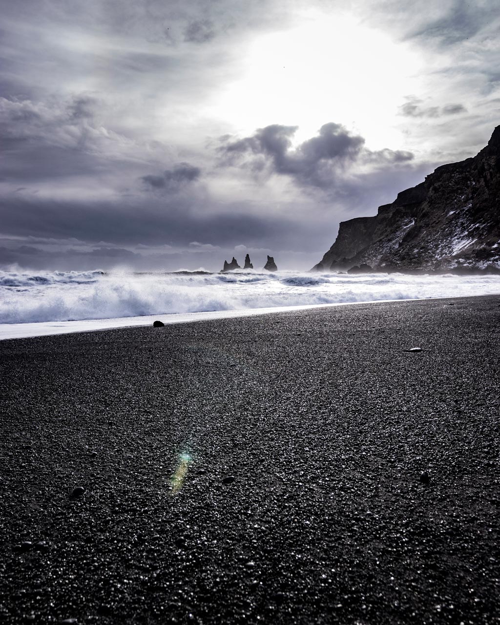 Waves break on black rock beach near the town of Vík, Iceland; the Reynisdrangar seastacks stand tall in the distance as dark spires in front of dark grey clouds.