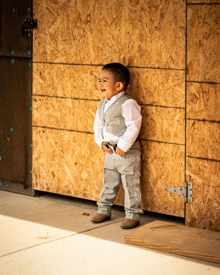 A 4-year old boy leans against a plywood door, dressed in a Charro suit, laughs at something out of frame.