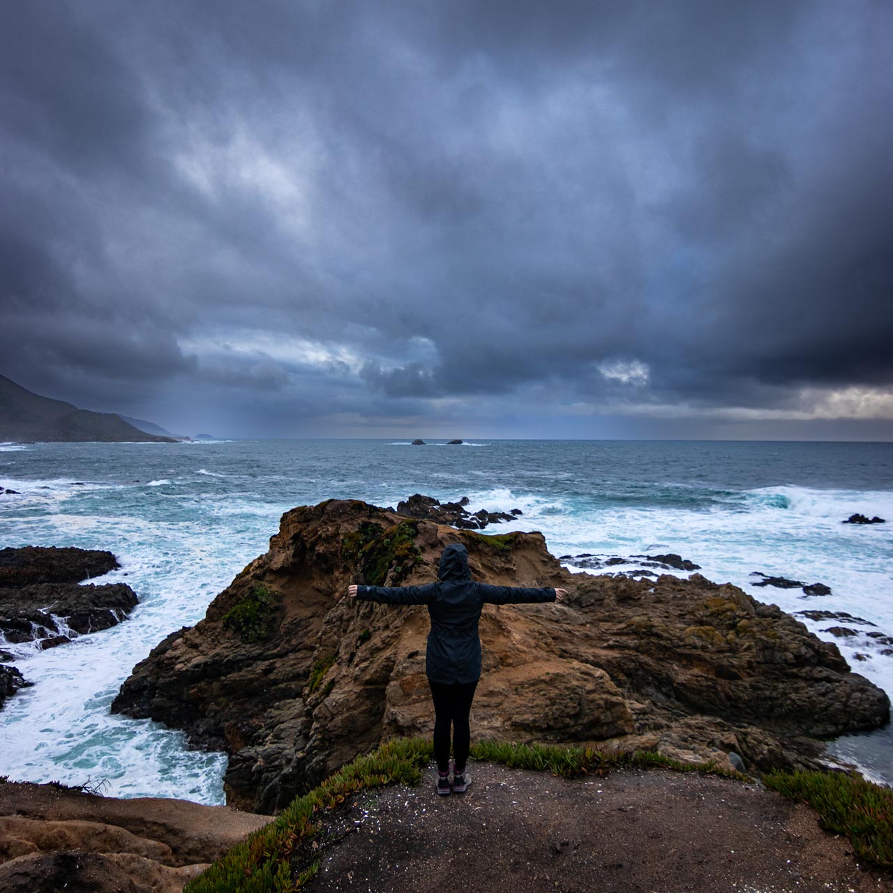 A woman stands with her arms spread open facing the Pacific Ocean in an early morning storm as the roiling clouds and turbulent waters rush the landscape she finds herself in.